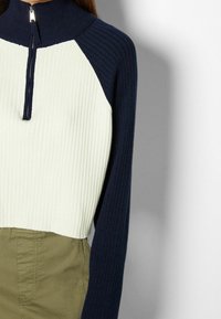 Ribbade zip-kragepullover i marinblått och krämfärgat, med kontrasterande raglansärmar och en strukturerad passform, kombinerad med olivgröna byxor.