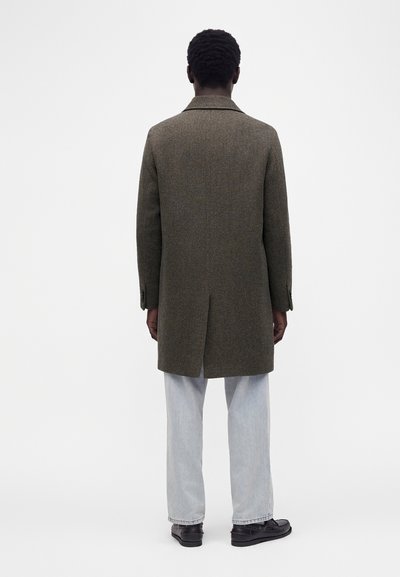 Homme vu de dos portant un pardessus marron texturé, un jean bleu clair et des mocassins noirs sur un fond blanc uni.