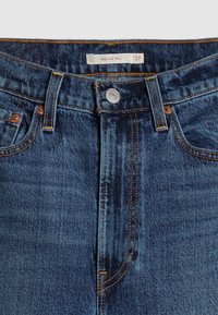 Gros plan sur la ceinture d'un jean en denim bleu avec un bouton métallique, des passants de ceinture, des poches avant et une étiquette intérieure portant l'inscription "RIBCAGE BELL 27".