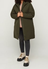 Zielona puchowa kurtka z kapturem, zapinana na zamek błyskawiczny, z dwoma przednimi kieszeniami. Do tego czarne dżinsy i beżowe buty na sznurowadła.