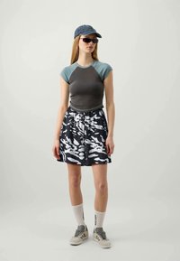Top ajustado de manga corta en gris y verde azulado, combinado con shorts de patrón en blanco y negro hasta la rodilla, zapatillas grises, calcetines blancos con patrón de texto y una gorra azul.