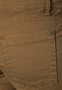 Beige bomullbyxor med en rund bakficka som har synliga sömmar och en metalldetalj i form av en nitar. Slät textur med en matt yta.