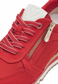 Chaussure de sport rouge présentant un mélange de matériaux lisses et perforés, accent de fermeture éclair dorée et lacets et languette contrastants en blanc.