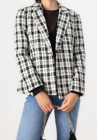 Blazer de tweed en patrón de cuadros blanco y negro, cierre de un botón, con solapas con muesca, combinado con denim y un bolso acentuado con cadena.