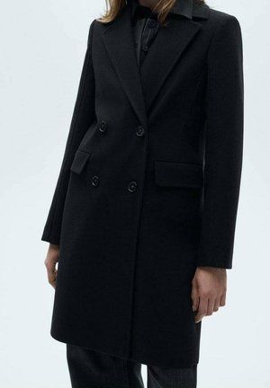 Manteau noir à double boutonnage avec larges revers et poches à rabat porté sur des vêtements sombres, présenté sur une personne debout des épaules jusqu'à mi-cuisse.