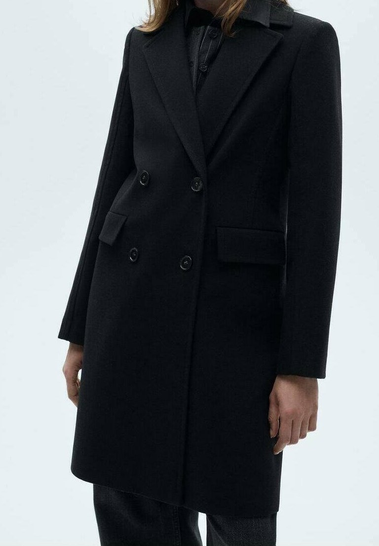 Manteau noir à double boutonnage avec larges revers et poches à rabat porté sur des vêtements sombres, présenté sur une personne debout des épaules jusqu'à mi-cuisse.