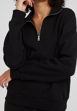 Osoba ubrana w czarny półgolf z długimi rękawami i cienki złoty naszyjnik, ręka dotykająca twarzy, ręce skrzyżowane.