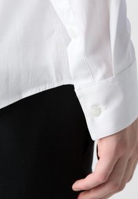 Weißes Dress-Shirt mit glatter Textur, Knopfmanschetten-Detail und abgerundetem Saum, kombiniert mit schwarzem Stoff darunter.