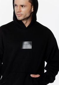 Czarna bluza z kapturem z prostokątną skórzaną naszywką z napisem "PILAWSKI". Materiał ma miękką strukturę, z przednią kieszenią typu kangur.