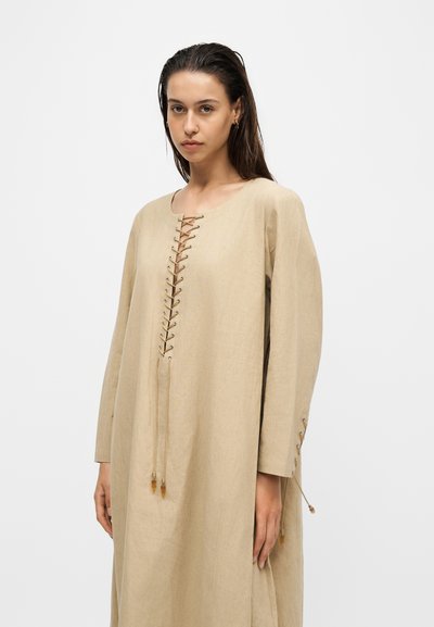 Femme aux cheveux foncés portant une longue robe beige avec des détails à lacets sur le devant et les manches, debout devant un fond clair uni.