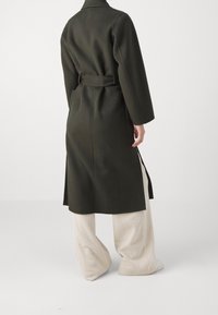 Abrigo de lana verde oscuro con cintura ceñida, mangas largas y corte recto, combinado con pantalones de pierna ancha de canalé en beige claro.