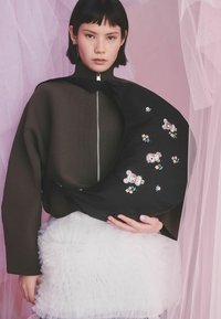 Lühikeste mustade juustega naine kannab tumedat lühemat tõmblukuga ülaosa ja valget volditud seelikut, hoides musta õlakotti, mis on kaunistatud kaisukaru ja lilledega.