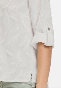 Blusa bianca con un motivo testurizzato a piume, maniche arrotolabili con dettagli a bottone e spacchi laterali per un tocco in più.