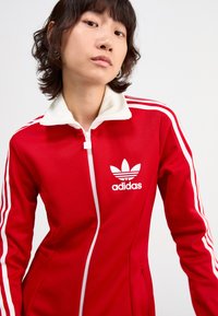Rood trainingspak met ritssluiting, gemaakt van polyester, met een witte kraag en zijkanten. Bevat een Adidas-logo op de borst. Glad textuur.
