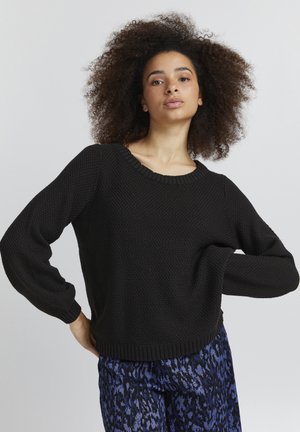 Pull en tricot noir avec un motif texturé, manches larges et col côtelé, associé à un pantalon bleu imprimé.
