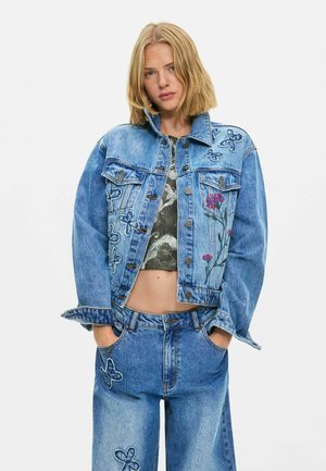 Junge Frau trägt eine blaue Jeansjacke und Jeans mit Schmetterlings- und Blumenstickerei, Hände in den Taschen, stehend vor einem schlichten weißen Hintergrund.