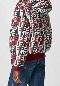 Vadderad jacka i kräm, röd och marinblå med logomönster. Har en huva, ribbade muddarna och en avslappnad passform. Jeansen i denim syns nedanför.