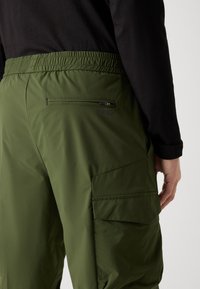 Grønne, lette cargo-bukser med elastisk liv, sidelomme og sikker lynlåslomme på bagsiden. Glat, mat tekstur.