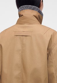 Personne aux cheveux noirs porte un manteau beige à col relevé avec un col intérieur en maille grise, vue de dos au niveau des épaules.
