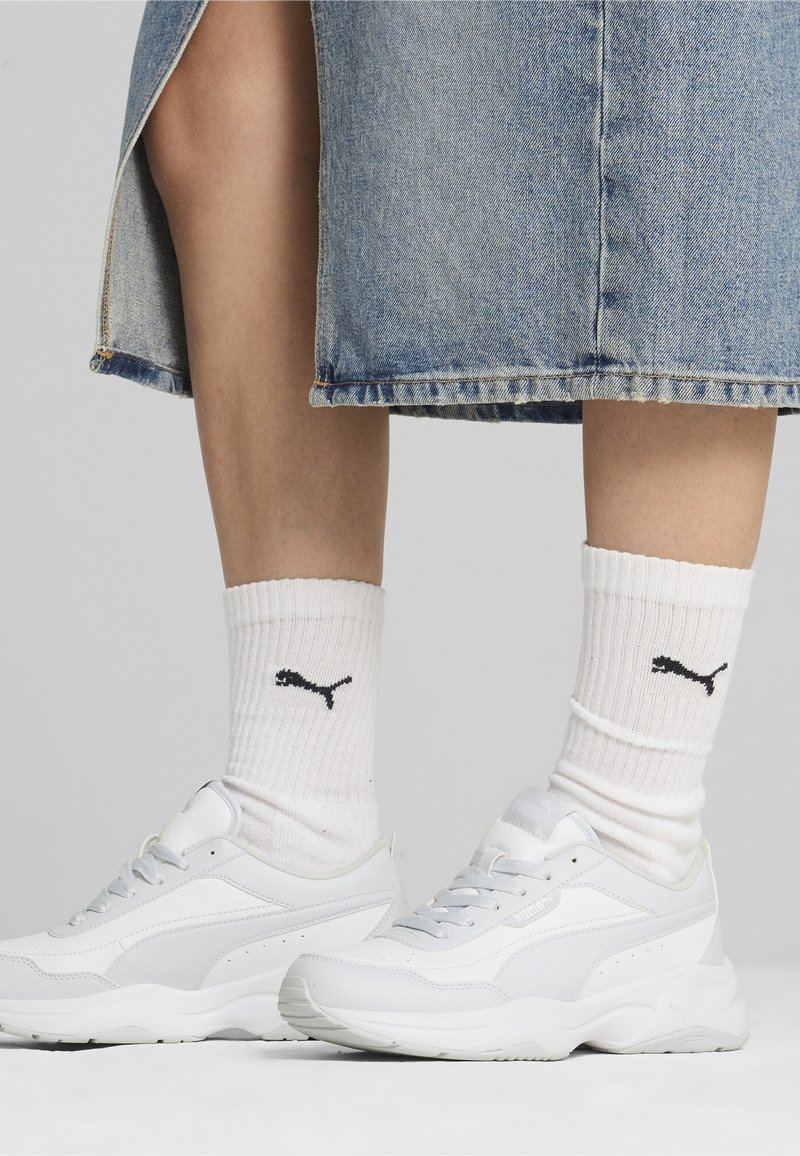 Jambes portant des chaussettes blanches Puma et des baskets blanches Puma associées à une jupe en jean bleue longueur genou sur fond gris.