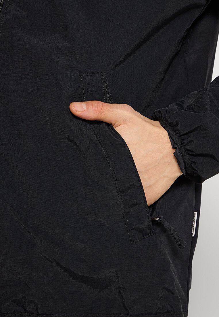 Giacca leggera nera con texture liscia, polsini elasticizzati e tasca laterale. Presenta una zip e un branding minimale sull'orlo.