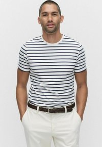 Gestreiftes T-Shirt in Weiß mit Marineblauen Streifen, kurzärmlig, mit Rundhalsausschnitt, kombiniert mit hellen Hosen und einem braunen geflochtenen Gürtel.