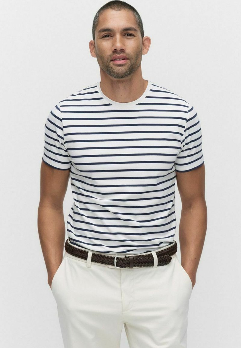 Gestreiftes T-Shirt in Weiß mit Marineblauen Streifen, kurzärmlig, mit Rundhalsausschnitt, kombiniert mit hellen Hosen und einem braunen geflochtenen Gürtel.
