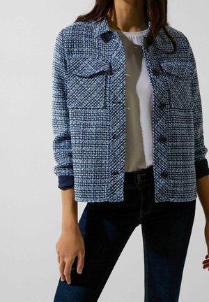 Kvinde iført en blå og hvid mønstret knap-op jakke over en hvid skjorte og mørke jeans mod en ensfarvet hvid baggrund.