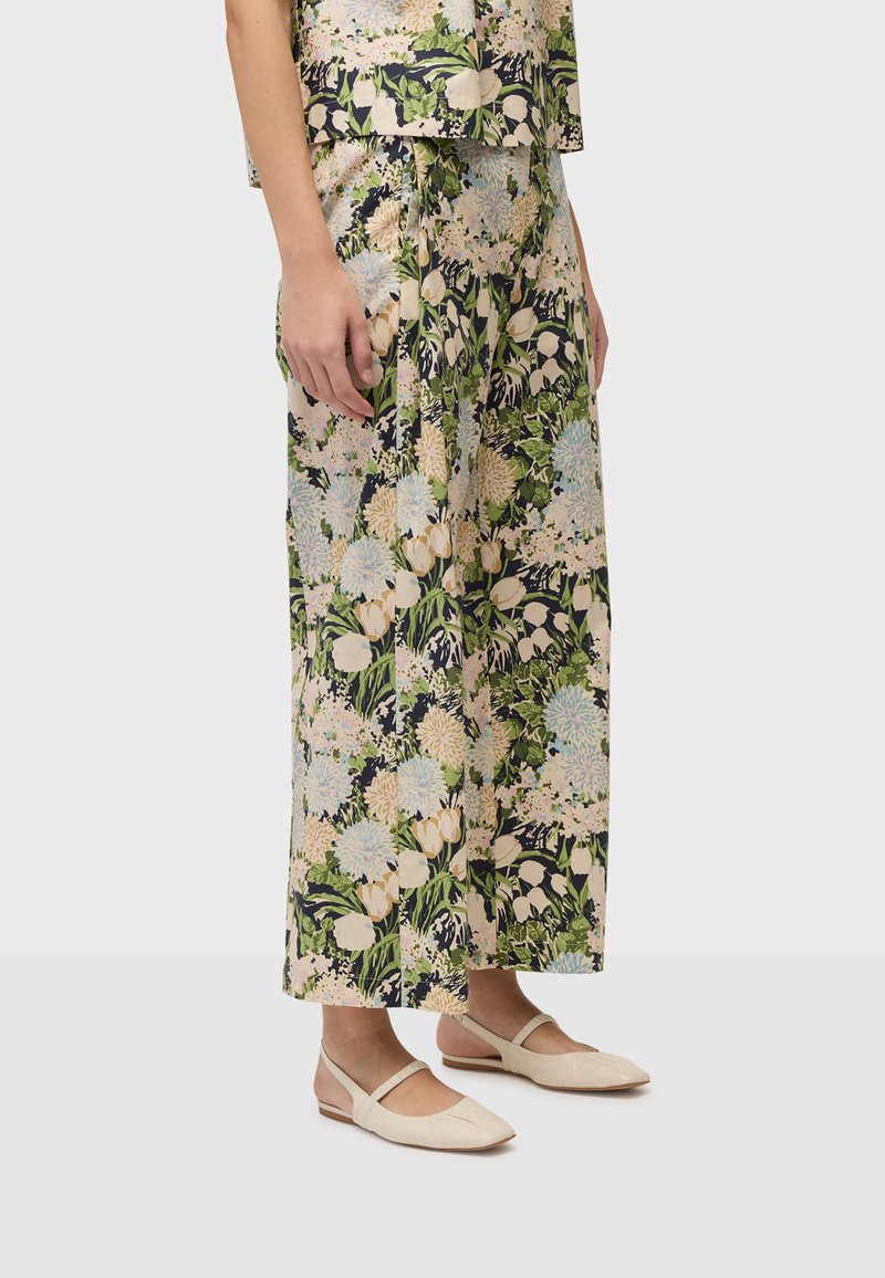 Femme portant un pantalon large à fleurs et un haut assorti avec des chaussures plates Mary Jane beige sur un fond gris clair.