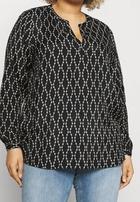 Sort bluse med et hvidt prik- og kæde-lignende mønster, med en udskåret halsudskæring og lange ærmer, lavet af et glat, let stof.