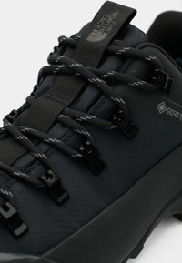 Črni pohodniški čevelj z teksturirano zgornjo površino, sivimi poudarki in vezalkami z plastičnimi kljukicami za varno pritrjevanje ter blagovno znamko GORE-TEX.