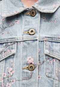 Φωτεινό μπλε τζιν μπουφάν με σχέδιο ροζ λουλουδιών, μεταλλικά κουμπιά, δύο τσέπες στο στήθος και εμφανείς λεπτομέρειες ραφής.