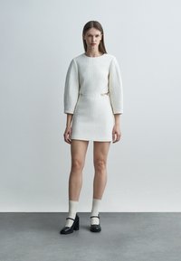 Žena v bielej minišaty s textúrou a nadýchanými trojštvrťovými rukávmi, doplnená bielymi ponožkami a čiernymi lodičkami Mary Jane, stojaca pri sivej stene.