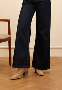 Beige leather ankle boots with a pointed toe and block heel, featuring subtle stitching details. Worn with dark wide-leg denim jeans.