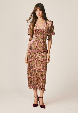 Mujer con vestido midi floral rosa y dorado con mangas abullonadas y tacones burdeos con pulsera en el tobillo, de pie frente a un fondo beige liso.