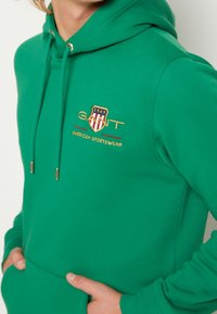 Vihreä huppari pehmeästä kankaasta, jossa kengurutasku, kultaiset nyörireiät ja kirjailtu logo punaisilla ja keltaisilla yksityiskohdilla.