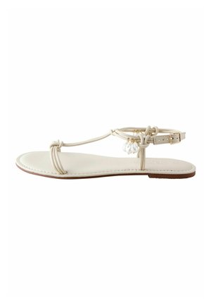 Beige flat sandal with thin straps, ankle buckle, and decorative small white beads dangling near the ankle strap.
