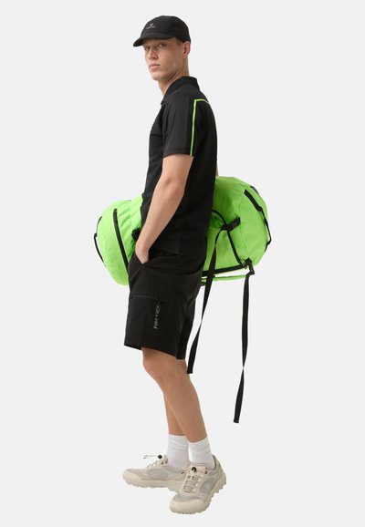 Černé polo s neonově zelenými detaily, černé šortky, bílé ponožky a béžové sneakers. Drží velkou, jasně zelenou sportovní tašku.