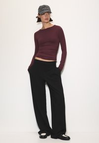 Egy bordó, testhez simuló, hosszú ujjú felsőt viselő nő, fekete, bő szárú nadrágban, fekete-fehér cipőben és egy szürke "New York City Brooklyn" sapkában.