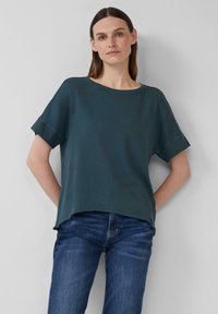 Teal kortearmige top met een relaxte pasvorm, ronde halslijn en licht gebogen zoom. Gecombineerd met blauwe jeans en gedragen tegen een neutrale achtergrond.
