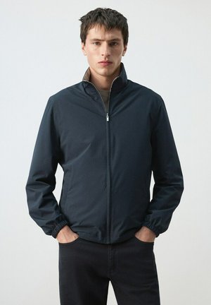 Joven con una chaqueta azul marino con cremallera y puños elásticos, de pie con las manos en los bolsillos frente a un fondo claro y sencillo.