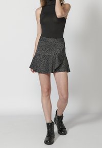 Femme portant un haut noir sans manches à col roulé, une mini-jupe imprimé léopard gris foncé, et des bottines noires à lacets, se tenant devant un fond blanc.