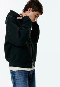 Felpa con zip nera in materiale felpato, con cappuccio, polsini a costine e uno strato interno bianco semplice, indossata con jeans in denim blu.