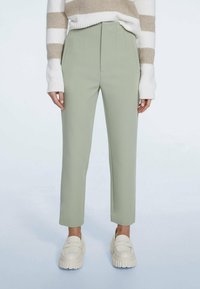 Pantalon slim vert sauge associé à des mocassins blancs épais et un pull rayé blanc et beige sur fond uni.