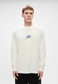 Λευκό κοντομάνικο μπλουζάκι με μακριά μανίκια, φτιαγμένο από μαλακό βαμβάκι, με λογότυπο στο στήθος σε μπλε και χρυσό χρώμα, συνδυασμένο με μαύρο παντελόνι.