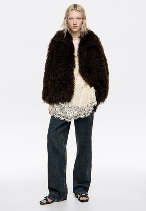 Blondynka w dużym, ciemnobrązowym płaszczu z futra sztucznego, z kremowym koronkowym topem, luźnych ciemnych dżinsach i plecionych sandałach na otwartym palcu stoi na tle białym.