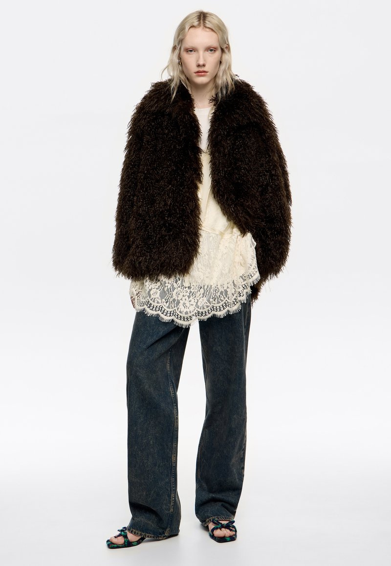 Mujer rubia que lleva un abrigo de piel sintética de gran tamaño en marrón oscuro, una blusa de encaje crema, unos vaqueros oscuros holgados y sandalias de dedo trenzadas, de pie contra un fondo blanco.