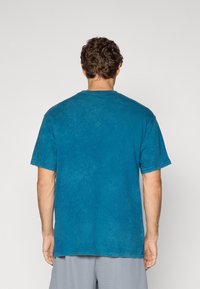 Mand med krøllet hår iført en vaskeblå kortærmet T-shirt og lysegrå shorts, vendt væk mod en ensfarvet lys baggrund.