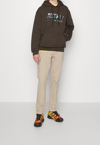 Felpa marrone con logo "Replay", pantaloni beige a gamba dritta e sneakers neri/arancioni con dettagli gialli. L'outfit è casual e funzionale.