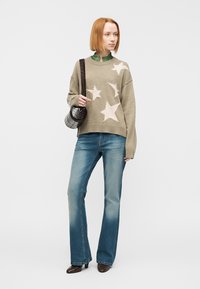 Πουλόβερ σε ελιά πράσινο με κρεμ μοτίβα αστεριών, συνδυασμένο με ανοιχτό μπλε καμπάνα τζιν. Το μοντέλο κρατά μια μικρή μαύρη τσάντα χιαστί. Ομαλή υφή.